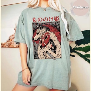 Op de afbeelding: Saliegroen T-shirt met een afbeelding van een vrouw, een wolf en de Japanse tekst "もののけ姫". De illustratie gebruikt rood, zwart en beige. Het shirt heeft korte mouwen en een losse pasvorm.