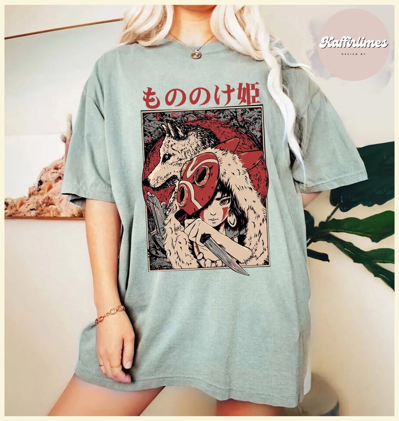Op de afbeelding: Saliegroen T-shirt met een afbeelding van een vrouw, een wolf en de Japanse tekst "もののけ姫". De illustratie gebruikt rood, zwart en beige. Het shirt heeft korte mouwen en een losse pasvorm.
