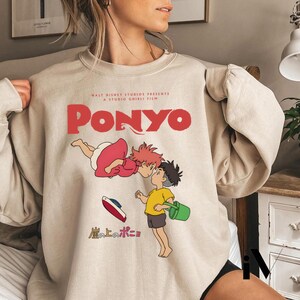 May include: Beige sweatshirt featuring a graphic from the animated film "Ponyo." The design includes the title "Ponyo" in red, along with illustrations of characters and Japanese text. The sweatshirt is a crew neck style.
