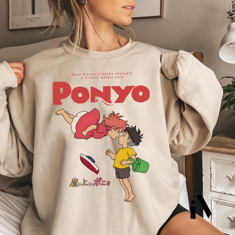 May include: Beige sweatshirt featuring a graphic from the animated film "Ponyo." The design includes the title "Ponyo" in red, along with illustrations of characters and Japanese text. The sweatshirt is a crew neck style.