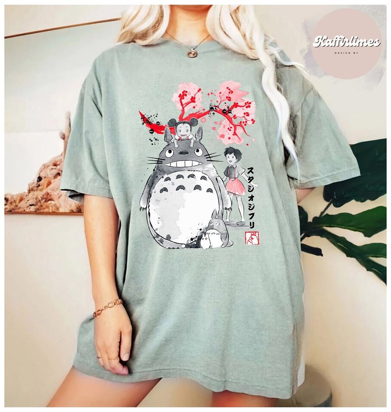 May include: Sage green t-shirt featuring a graphic of Totoro and other characters from the animated film My Neighbor Totoro. The design includes a cherry blossom branch and Japanese text. The shirt is a relaxed fit.