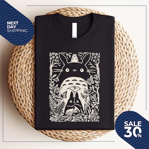 May include: Black t-shirt featuring a white graphic of a forest scene with the characters from the animated film, My Neighbor Totoro. The design includes Totoro, butterflies, and other forest elements. The shirt is folded on a woven surface.