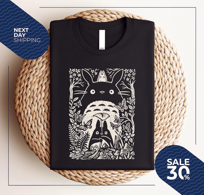 May include: Black t-shirt featuring a white graphic of a forest scene with the characters from the animated film, My Neighbor Totoro. The design includes Totoro, butterflies, and other forest elements. The shirt is folded on a woven surface.