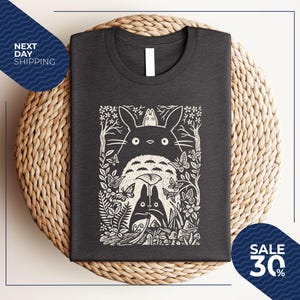 May include: Dark gray t-shirt featuring a white graphic of a forest scene with a large, round creature and smaller figures. The design includes trees, butterflies, and foliage. The shirt is folded on a woven surface.