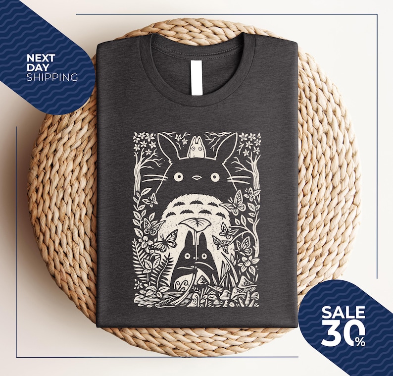 May include: Dark gray t-shirt featuring a white graphic of a forest scene with a large, round creature and smaller figures. The design includes trees, butterflies, and foliage. The shirt is folded on a woven surface.