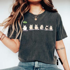 Könnte beinhalten: Dunkelgraues T-Shirt mit einer Reihe von Cartoon-Figuren mit unterschiedlichen Gesichtsausdrücken. Die Figuren sind weiß, beige und grau, mit schwarzen Augen und Details. Ein grünes Blatt schwebt über der ersten Figur.