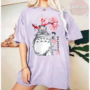 May include: A light purple t-shirt featuring a graphic of characters from the animated film "My Neighbor Totoro." The design includes Totoro, a girl, and cherry blossoms. The text "スタジオジブリ" is also visible.