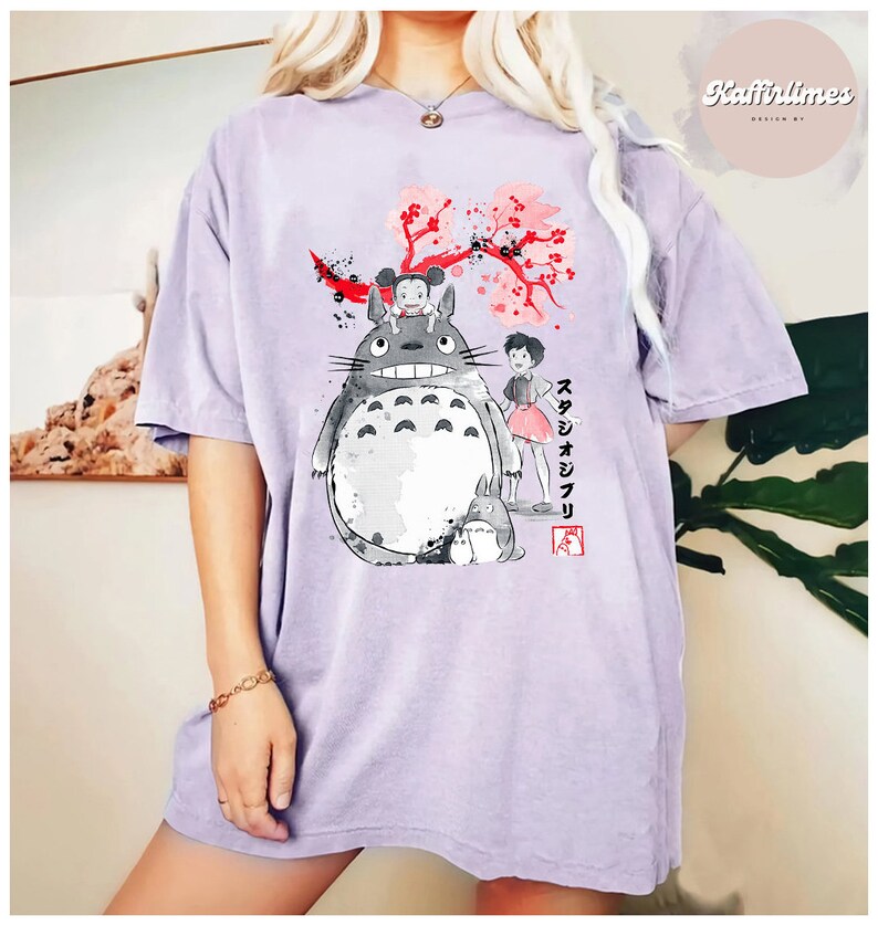 May include: A light purple t-shirt featuring a graphic of characters from the animated film "My Neighbor Totoro." The design includes Totoro, a girl, and cherry blossoms. The text "スタジオジブリ" is also visible.