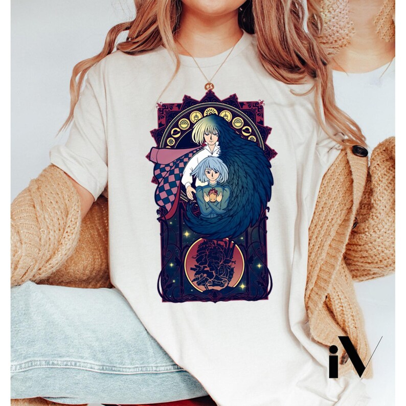 May include: Cream-colored t-shirt featuring a detailed illustration of two anime characters within a decorative frame. The artwork showcases a blonde character with a pink cape and a blue-haired character. The design is in shades of blue, purple, and gold.
