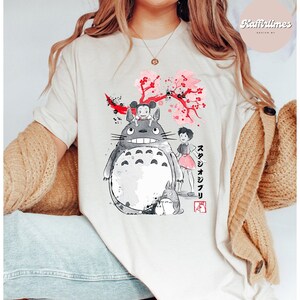 May include: Cream-colored t-shirt featuring a graphic of Totoro and other characters from the animated film My Neighbor Totoro. The design includes a large gray Totoro, a smaller Totoro, and a girl in a pink dress, with Japanese text and cherry blossoms.