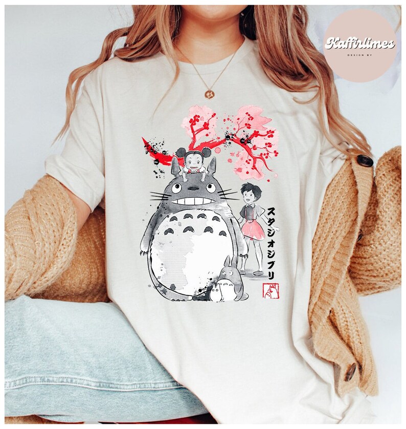 May include: Cream-colored t-shirt featuring a graphic of Totoro and other characters from the animated film My Neighbor Totoro. The design includes a large gray Totoro, a smaller Totoro, and a girl in a pink dress, with Japanese text and cherry blossoms.
