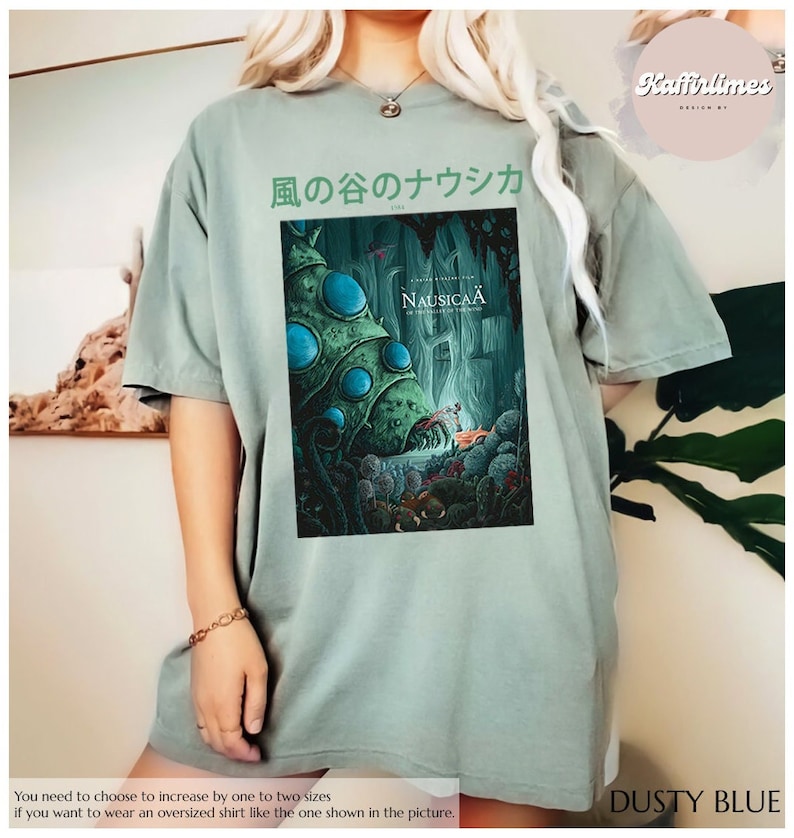 Może przedstawiać: Oversize'owa koszulka w kolorze przydymionego błękitu z grafiką z filmu "Nausica&auml; z Doliny Wiatru". Projekt zawiera japoński tekst i kolorową ilustrację. Koszulka ma luźny kr&oacute;j.