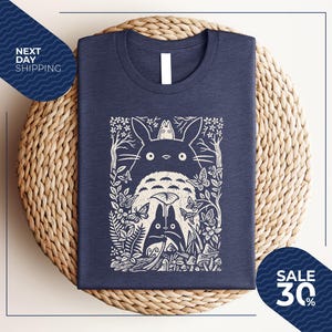 May include: Navy blue t-shirt featuring a white graphic of a forest scene with the characters from the animated film, My Neighbor Totoro. The design includes Totoro, butterflies, and foliage. The shirt is displayed on a woven surface.