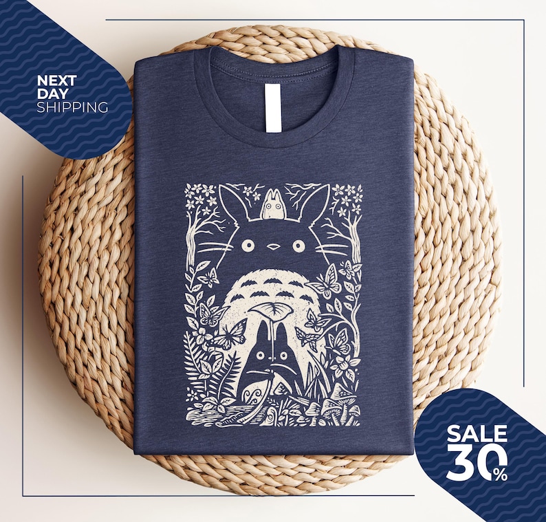 May include: Navy blue t-shirt featuring a white graphic of a forest scene with the characters from the animated film, My Neighbor Totoro. The design includes Totoro, butterflies, and foliage. The shirt is displayed on a woven surface.