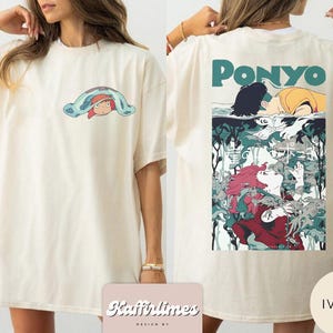May include: Cream-coloured t-shirt with a front graphic of a cartoon character under a blue blanket. The back of the shirt features the word "Ponyo" in teal above a colourful anime-style illustration. The shirt has a relaxed, oversized fit.