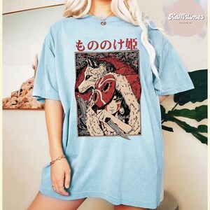 Op de afbeelding: Lichtblauw T-shirt met een afbeelding van een vrouw, een wolf en Japanse tekst. De afbeelding is rood, wit en zwart. Het shirt heeft een relaxte, oversized pasvorm.