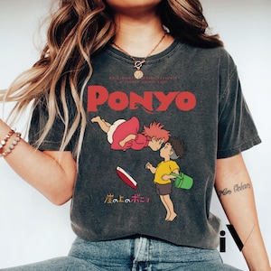 May include: Dark gray t-shirt featuring the word "PONYO" in red, with an illustration of characters from the animated film. The design includes a girl in a pink dress and a boy in a yellow shirt, with Japanese text below.
