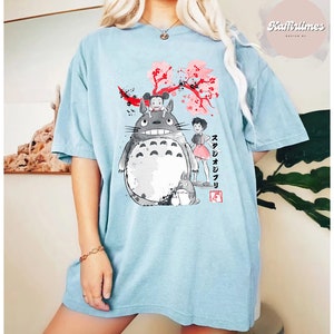 May include: Light blue t-shirt featuring a graphic of characters from the animated film, My Neighbor Totoro. The design includes Totoro, two girls, and cherry blossoms. Japanese text is also visible.