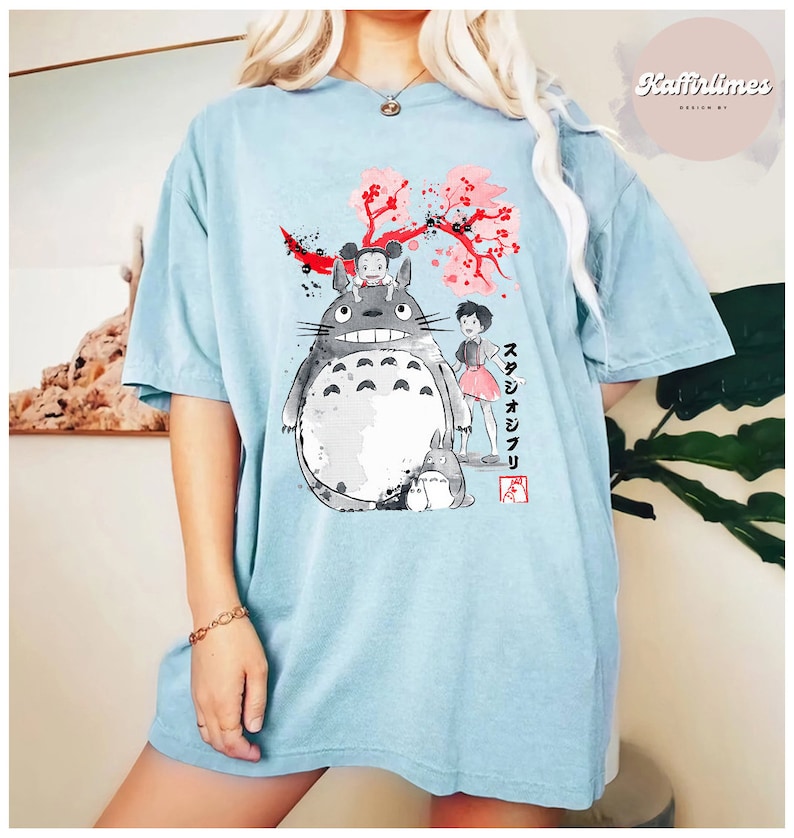 May include: Light blue t-shirt featuring a graphic of characters from the animated film, My Neighbor Totoro. The design includes Totoro, two girls, and cherry blossoms. Japanese text is also visible.