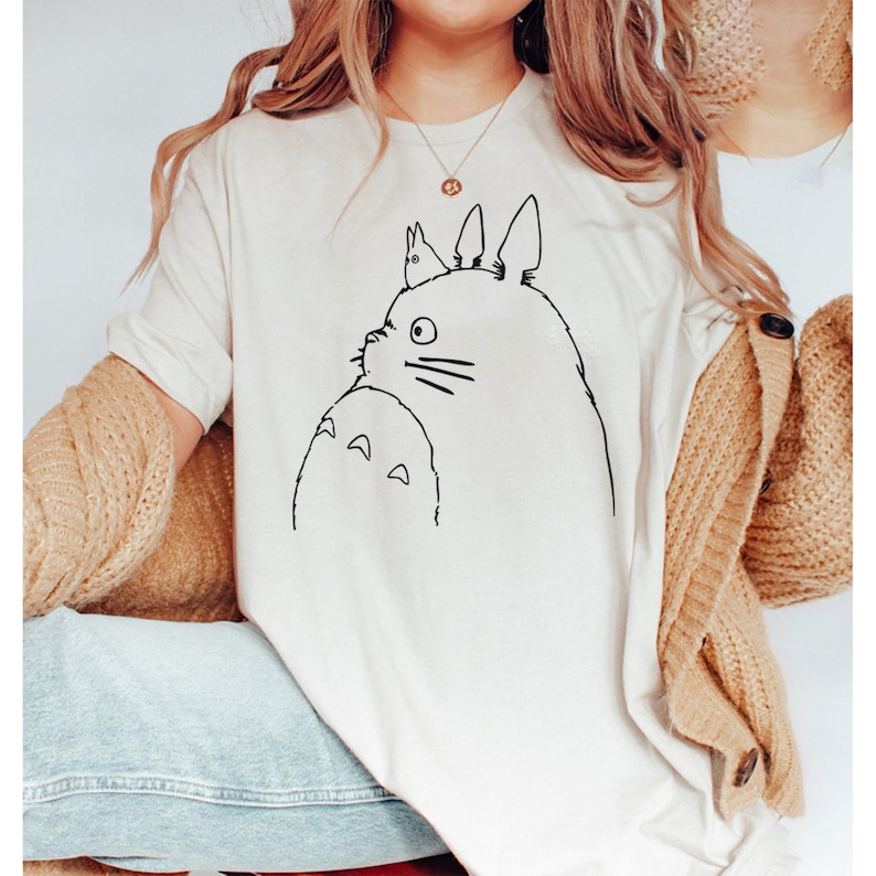 May include: Cream-colored t-shirt featuring a black line drawing of a Totoro character. The design includes the character's face, whiskers, and belly. The shirt is a casual, short-sleeved style.