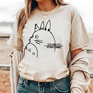 May include: Beige t-shirt featuring a black line drawing of a cartoon character with large ears and the text "STUDIO GHIBLI" in black. The shirt has short sleeves and a crew neck.