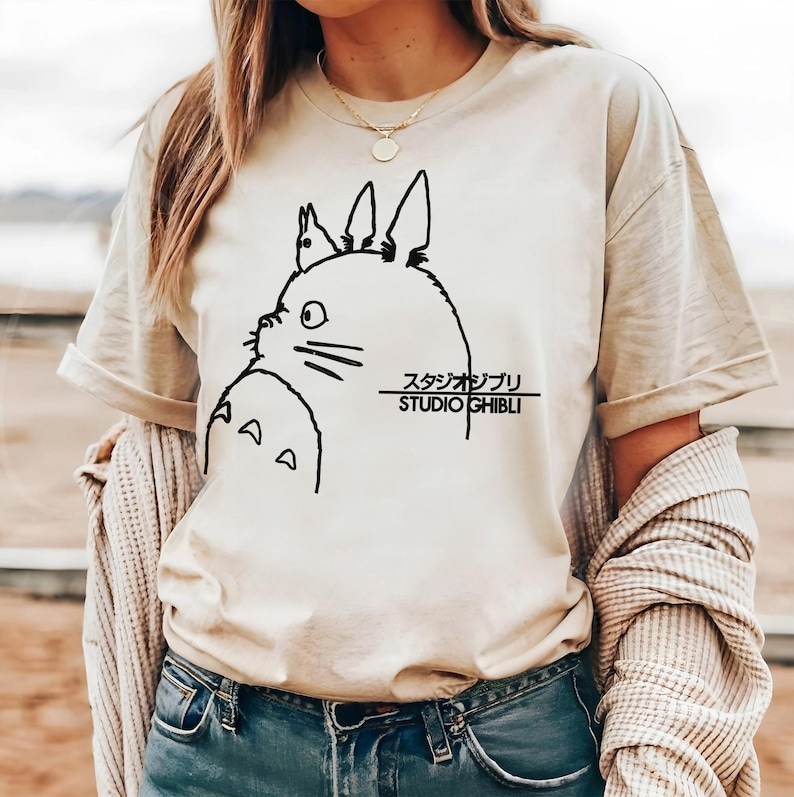 May include: Beige t-shirt featuring a black line drawing of a cartoon character with large ears and the text "STUDIO GHIBLI" in black. The shirt has short sleeves and a crew neck.