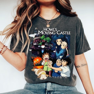 May include: Dark grey t-shirt with a colourful graphic from the animated film "Howl's Moving Castle." The design includes characters and the film's title. The shirt is a crew neck style.