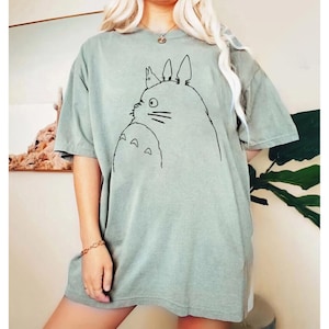 May include: Sage green t-shirt featuring a black line drawing of a cartoon character. The illustration depicts a large, round creature with pointed ears and a smaller version of itself. The shirt has short sleeves and a relaxed fit.
