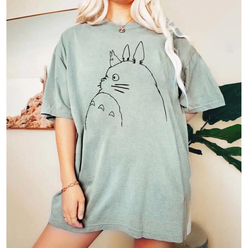 May include: Sage green t-shirt featuring a black line drawing of a cartoon character. The illustration depicts a large, round creature with pointed ears and a smaller version of itself. The shirt has short sleeves and a relaxed fit.