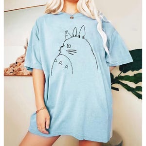May include: Light blue t-shirt featuring a black line drawing of a cartoon character. The design includes a large, round creature with pointed ears and a smaller creature below. The shirt has short sleeves and a relaxed fit.