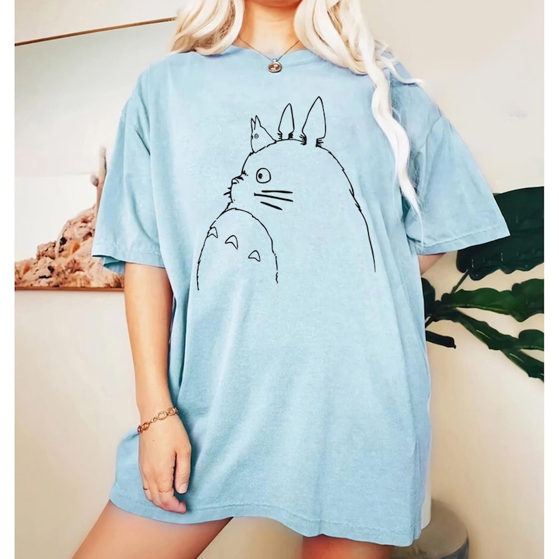 May include: Light blue t-shirt featuring a black line drawing of a cartoon character. The design includes a large, round creature with pointed ears and a smaller creature below. The shirt has short sleeves and a relaxed fit.