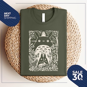 May include: Olive green t-shirt featuring a white graphic of a forest scene with characters. The design includes a large, round creature with a smaller one on top, surrounded by trees, butterflies, and foliage. The shirt is folded on a woven surface.