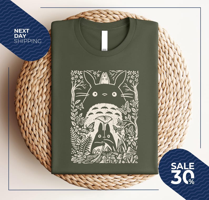 May include: Olive green t-shirt featuring a white graphic of a forest scene with characters. The design includes a large, round creature with a smaller one on top, surrounded by trees, butterflies, and foliage. The shirt is folded on a woven surface.
