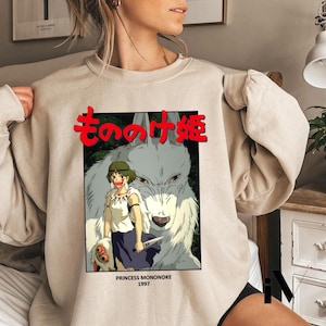 May include: Beige sweatshirt featuring a graphic from the animated film "Princess Mononoke." The design includes Japanese text, a character illustration, and the year 1997. The artwork depicts a woman with a wolf.