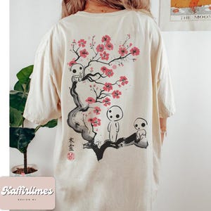 May include: Cream-colored t-shirt featuring a Japanese-inspired design. The back of the shirt displays a black and red illustration of a flowering tree with spirit-like figures. The text "木霊" is also visible.