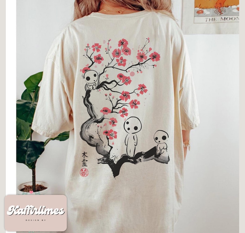 May include: Cream-colored t-shirt featuring a Japanese-inspired design. The back of the shirt displays a black and red illustration of a flowering tree with spirit-like figures. The text "木霊" is also visible.