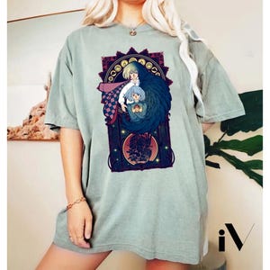 May include: Sage green t-shirt featuring a detailed anime-style illustration. The graphic showcases three figures within an ornate, dark red and gold frame. The central figures are a blonde-haired person, a blue-haired person, and a figure with dark blue wings.