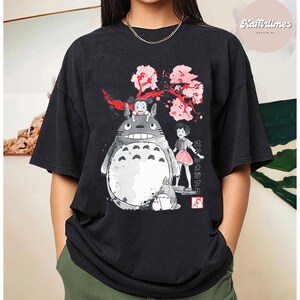 May include: Black t-shirt featuring a graphic of Totoro and other characters from the animated film My Neighbor Totoro. The design includes a large gray Totoro, a small child, and a pink cherry blossom branch. The text "スタジオジブリ" is also visible.