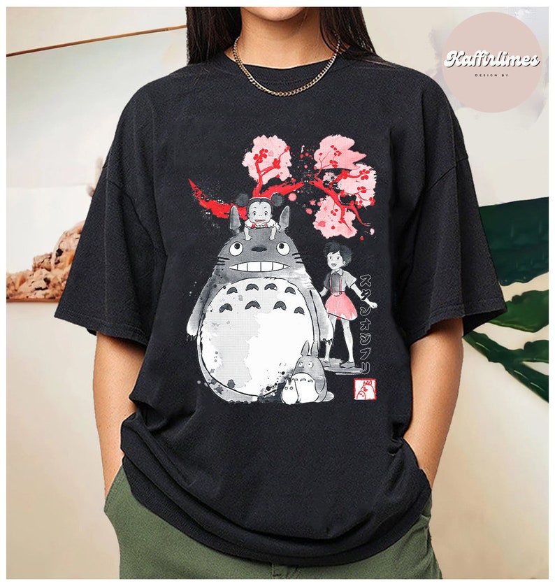 May include: Black t-shirt featuring a graphic of Totoro and other characters from the animated film My Neighbor Totoro. The design includes a large gray Totoro, a small child, and a pink cherry blossom branch. The text "スタジオジブリ" is also visible.