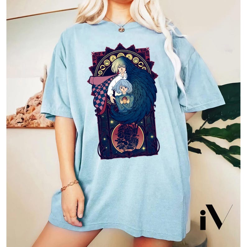 May include: Light blue t-shirt featuring a colorful graphic from an animated film. The graphic includes two characters with long hair, one blonde and one blue, within a decorative frame. The design is centered on the shirt.