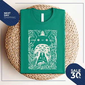May include: A green t-shirt featuring a white graphic of a forest scene with characters. The graphic includes a large, fluffy creature with large eyes, smaller creatures, butterflies, and foliage. The shirt is folded on a woven mat.