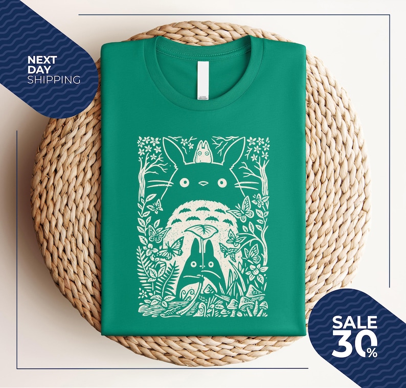 May include: A green t-shirt featuring a white graphic of a forest scene with characters. The graphic includes a large, fluffy creature with large eyes, smaller creatures, butterflies, and foliage. The shirt is folded on a woven mat.
