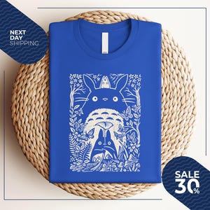 May include: Royal blue t-shirt featuring a white graphic of a forest scene with characters. The design includes a large, round creature with two smaller figures, butterflies, and foliage. The shirt is folded on a woven surface.