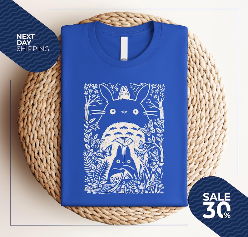 May include: Royal blue t-shirt featuring a white graphic of a forest scene with characters. The design includes a large, round creature with two smaller figures, butterflies, and foliage. The shirt is folded on a woven surface.