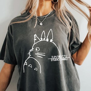 May include: Dark gray t-shirt featuring a white outline of a cartoon character from Studio Ghibli. The text "スタジオジブリ STUDIO GHIBLI" is printed in white below the character. The shirt has short sleeves.