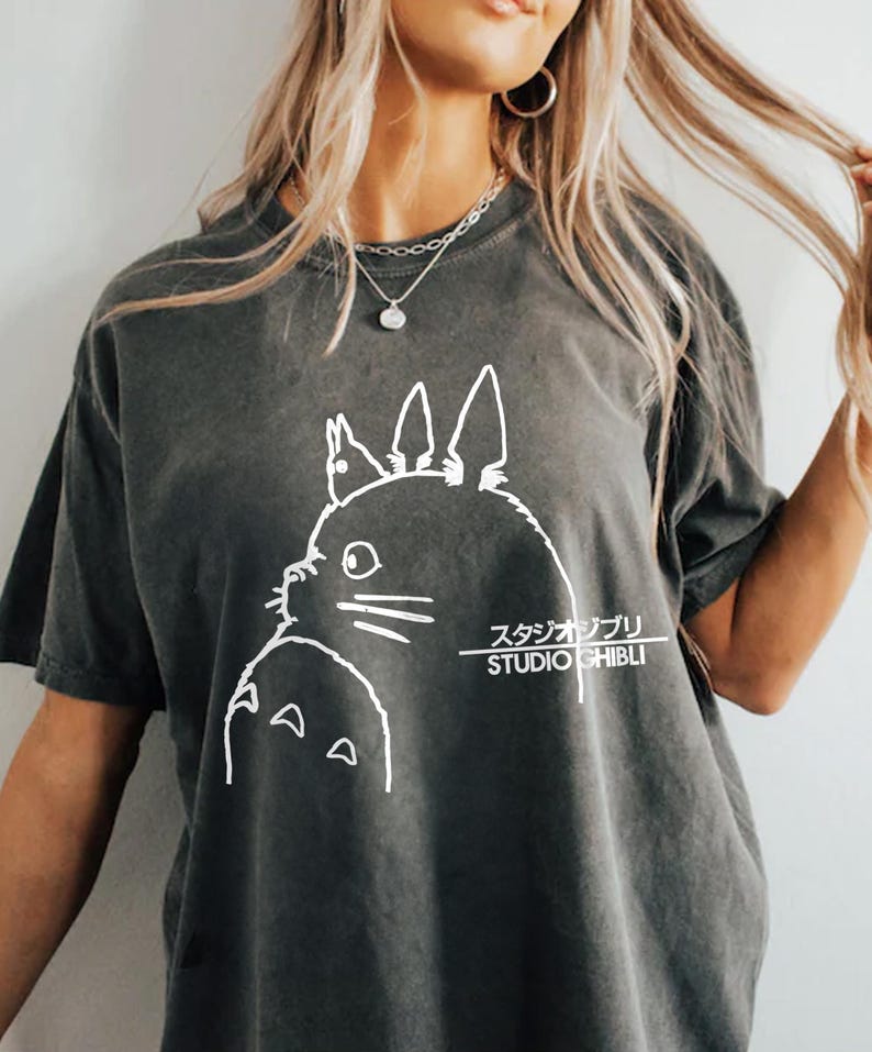 May include: Dark gray t-shirt featuring a white outline of a cartoon character from Studio Ghibli. The text "スタジオジブリ STUDIO GHIBLI" is printed in white below the character. The shirt has short sleeves.