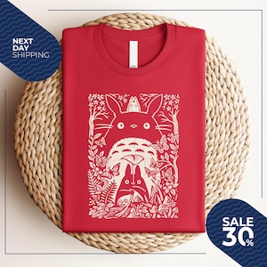 May include: Red t-shirt featuring a white graphic of characters from a popular animated film. The design includes a large, fluffy creature with a smaller one on top, surrounded by butterflies, trees, and foliage. The shirt is folded neatly on a woven surface.