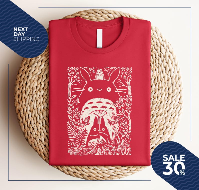 May include: Red t-shirt featuring a white graphic of characters from a popular animated film. The design includes a large, fluffy creature with a smaller one on top, surrounded by butterflies, trees, and foliage. The shirt is folded neatly on a woven surface.