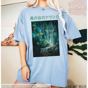 Może przedstawiać: Jasnoniebieska koszulka oversize z grafiką z filmu animowanego "Nausica&auml; z Doliny Wiatru". Projekt zawiera japoński tekst i kolorową ilustrację. Koszulka jest jasnoniebieska.