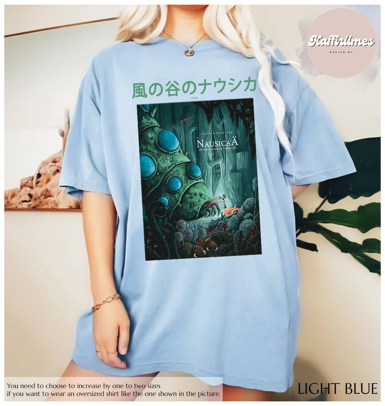 Może przedstawiać: Jasnoniebieska koszulka oversize z grafiką z filmu animowanego "Nausica&auml; z Doliny Wiatru". Projekt zawiera japoński tekst i kolorową ilustrację. Koszulka jest jasnoniebieska.